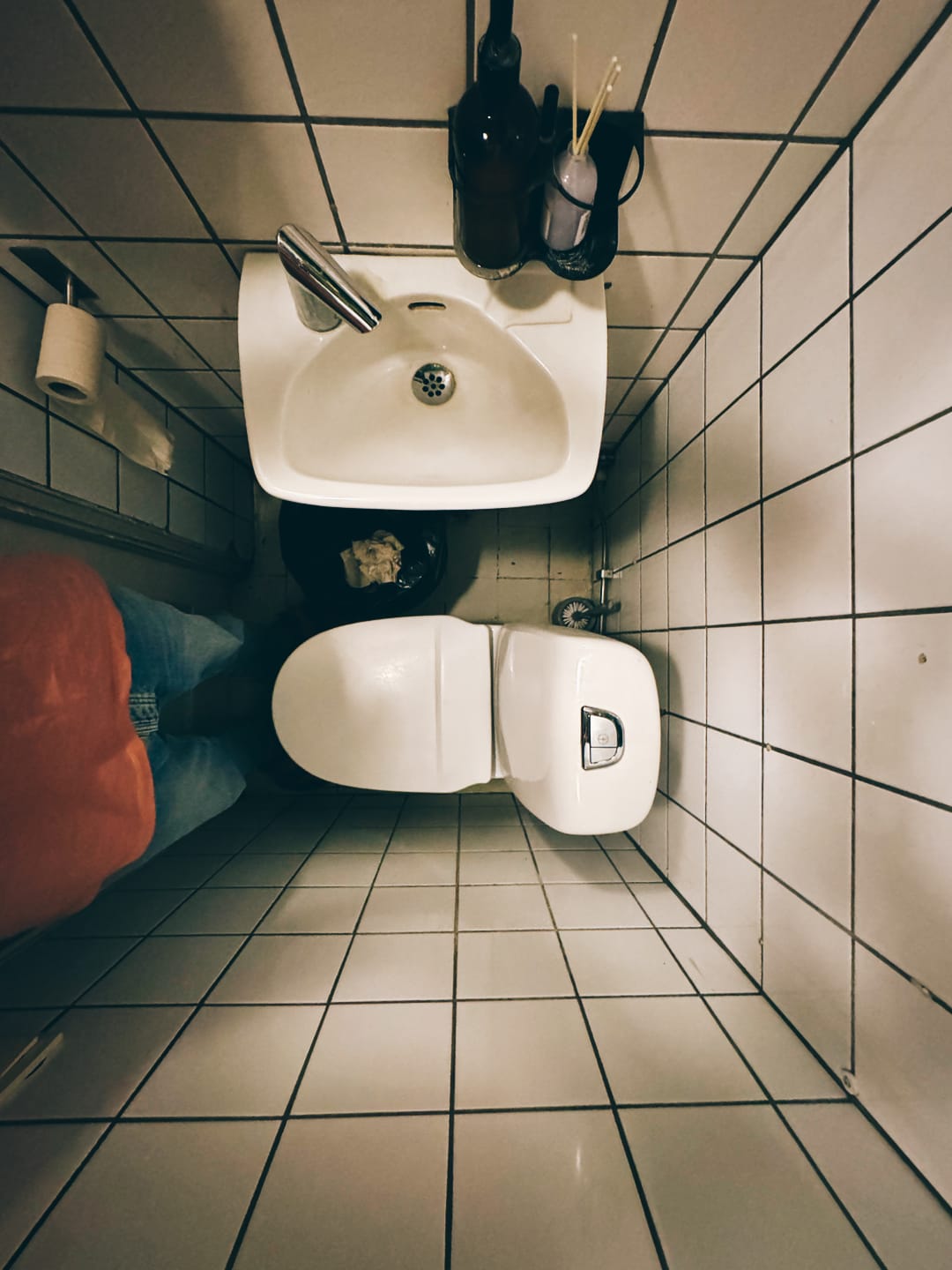 Fisheye view looking straight down into a tiny square bathroom with white ceramic tiles on the floor and walls. A toilet, a sink, and yours truly compete for the available square footage.