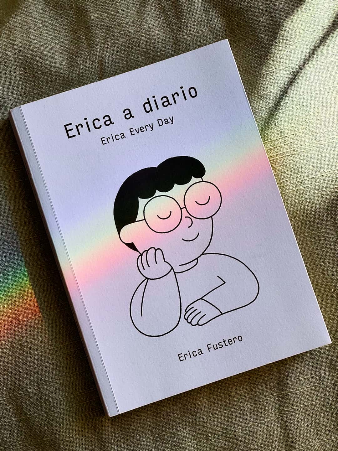The book rests on a pillow, its cover featuring a simple self-portrait drawing of Erica, at peace with shut eyes. When I snapped the photo, a rainbow happened to line up perfectly with her glasses—it looks like she&rsquo;s sunbathing.