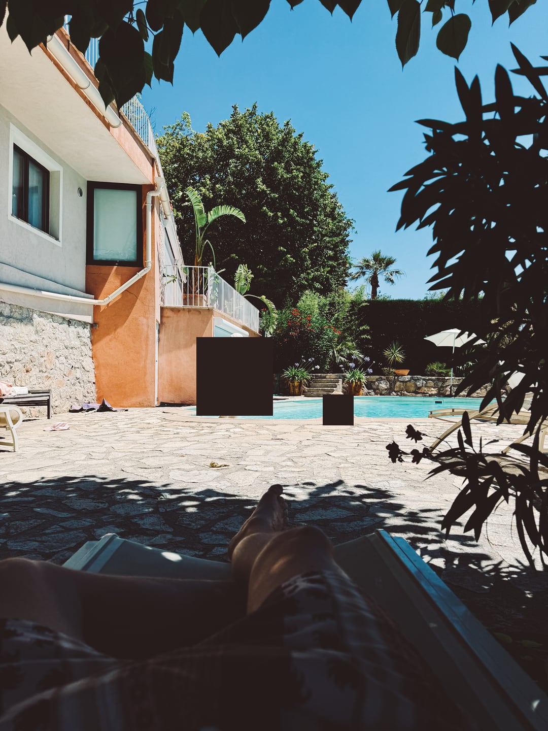 From a sunbed in the shade, with my legs visible in the foreground and the pool stretching out ahead on a perfect summer day. Two black rectangles partially obscure the image.