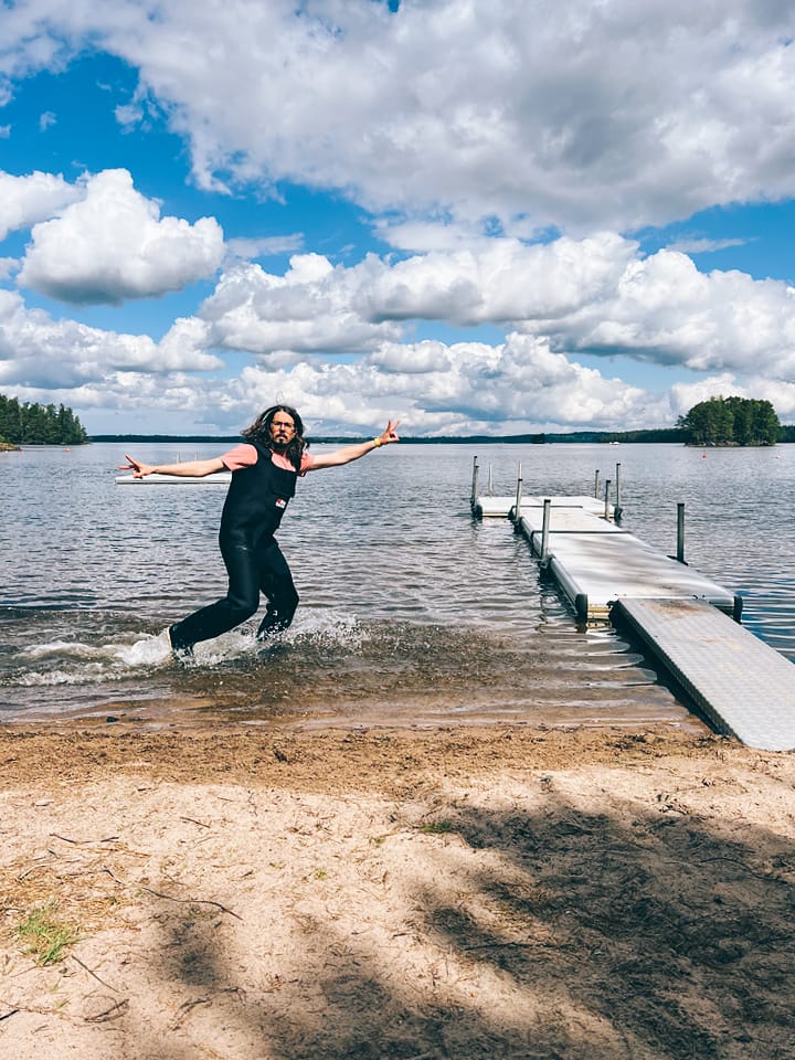 A serene lakeside view of a floating dock stretching out from the beach into the water. A middle-aged man in waders completely ruins the peaceful vibe by sprinting through the shallows with a dead-serious expression while throwing double peace signs.
