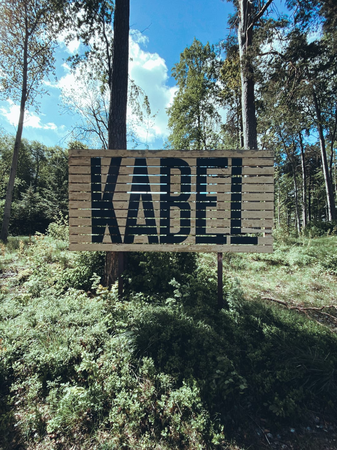 There is a big wooden sign surrounded by trees and greenness, seemingly in the middle of the forest. The word KABEL is written using giant, black letters in all caps.