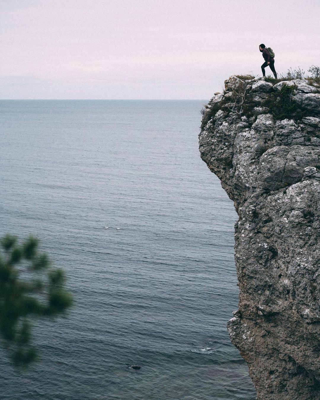 A woman is standing at the edge of a cliff. Down below, the wind ruffles the water. The sky is overcast.