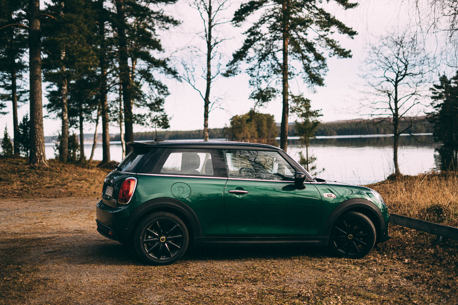 The profile of a green, three-door car. It stands parked outdoors; there's a lake in the background, and it looks like autumn.