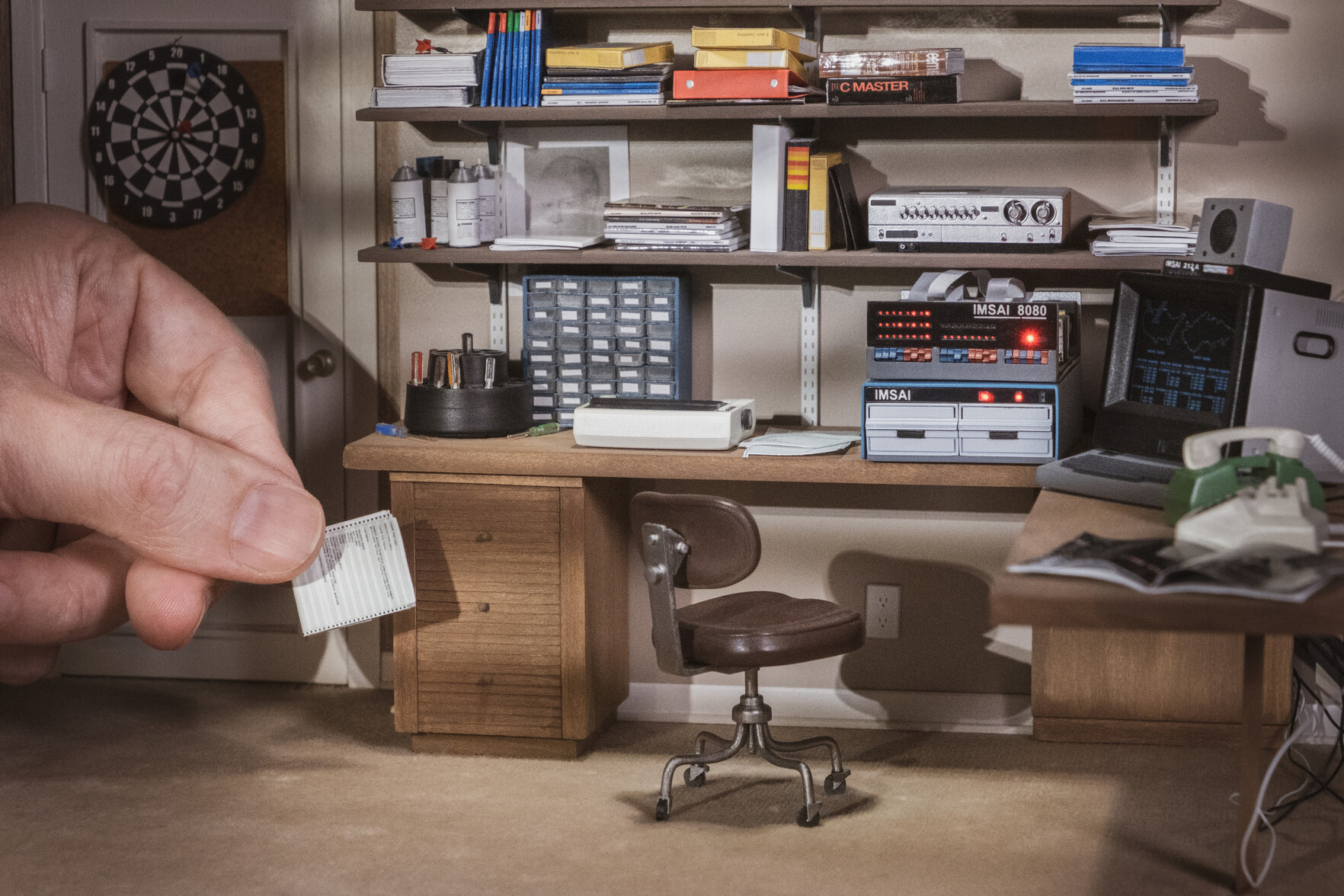 Miniature 1:12 scale model of David Lightman bedroom from the movie Wargames from 1983. A normal sized hand is seen entering the frame of the picture from the left, holding a miniature printout paper.
