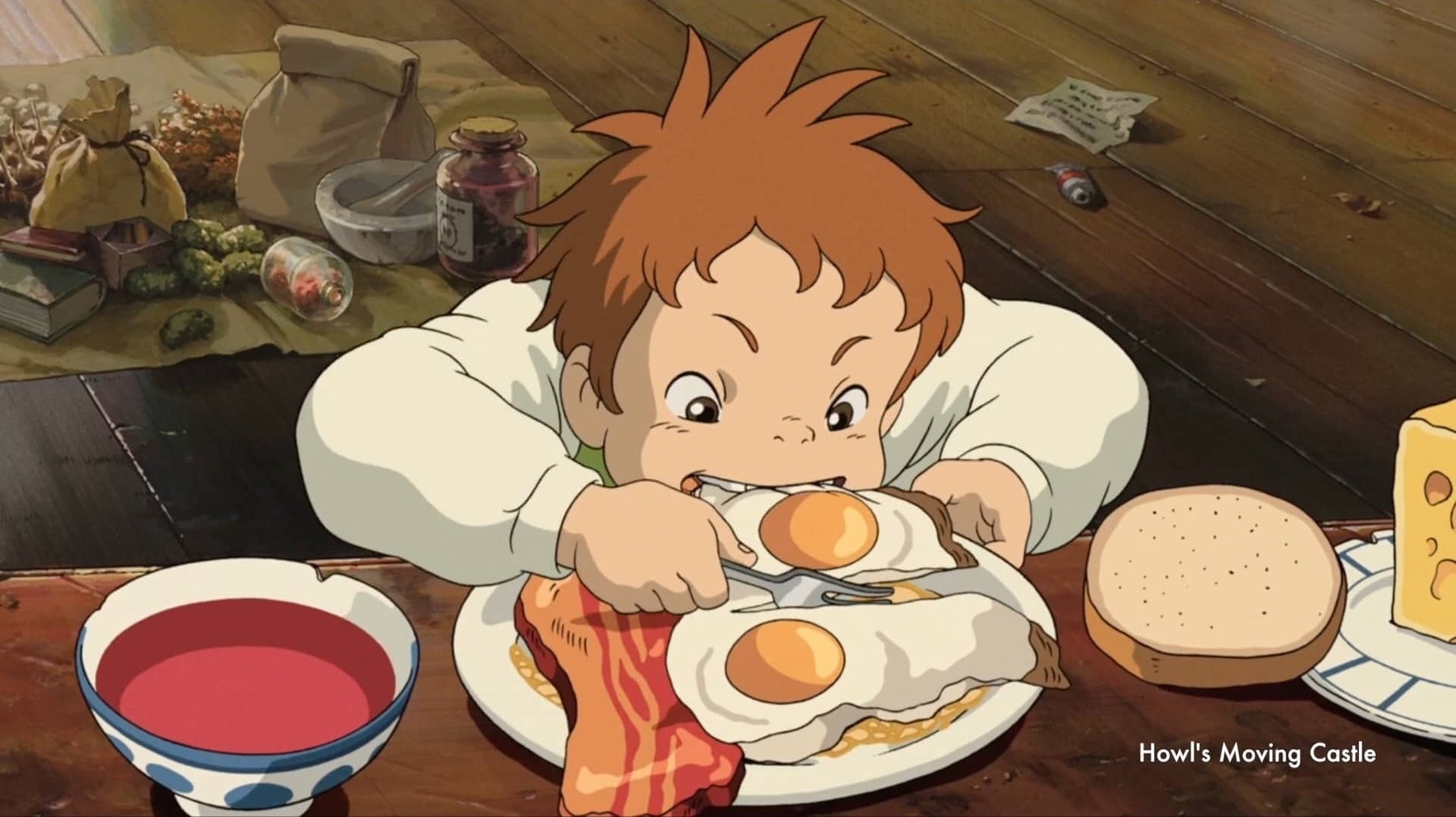 Single frame from the anime Howl’s Moving Castle. The table is set with food: soup, bread, cheese, egg, and meat. A boy gobbles the eggs with enthusiasm.