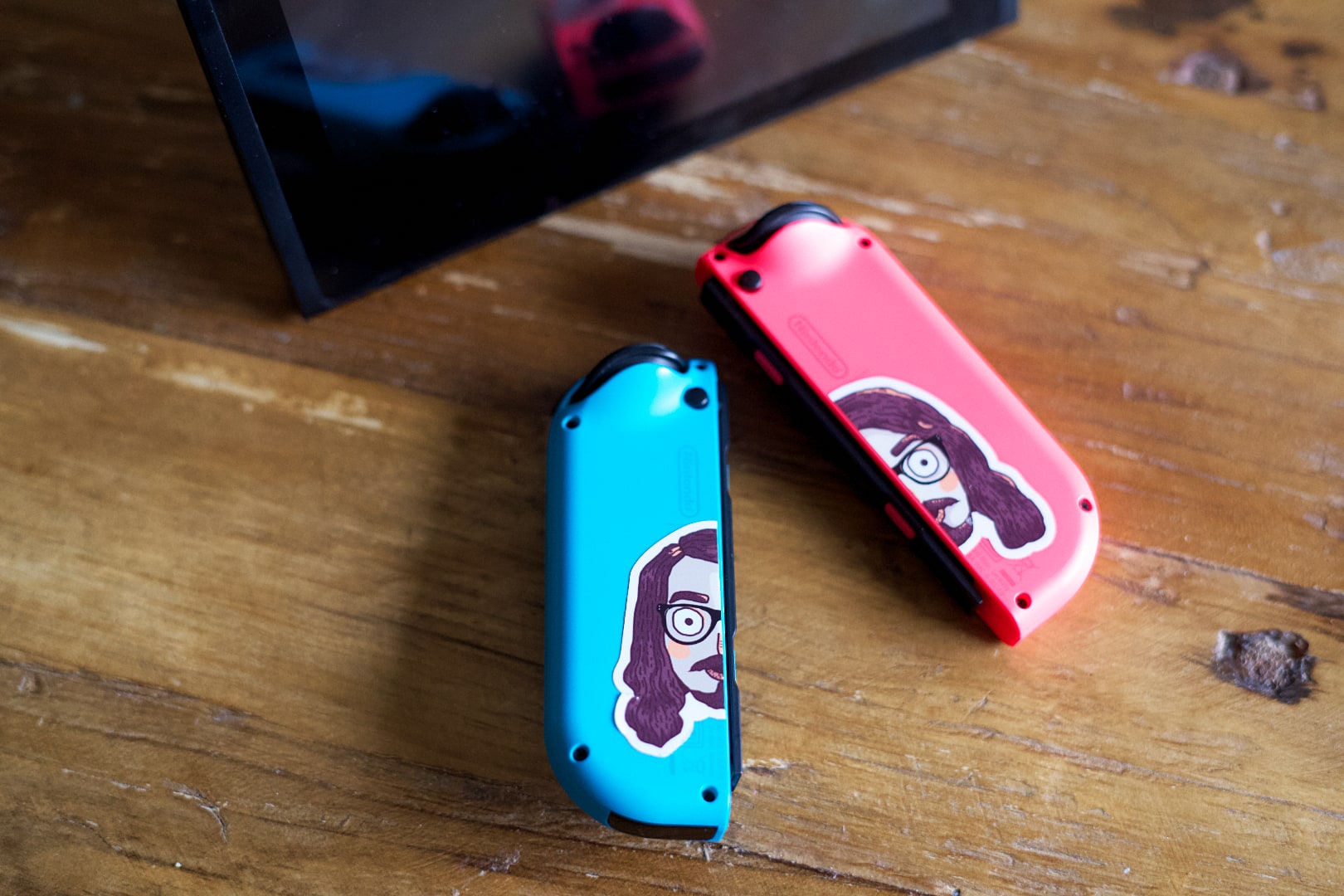 A pair of Joy-Con controllers with a sticker on the back featuring the head of a guy with long, brown hair, glasses, and a beard. The sticker is split in half, one for each controller.