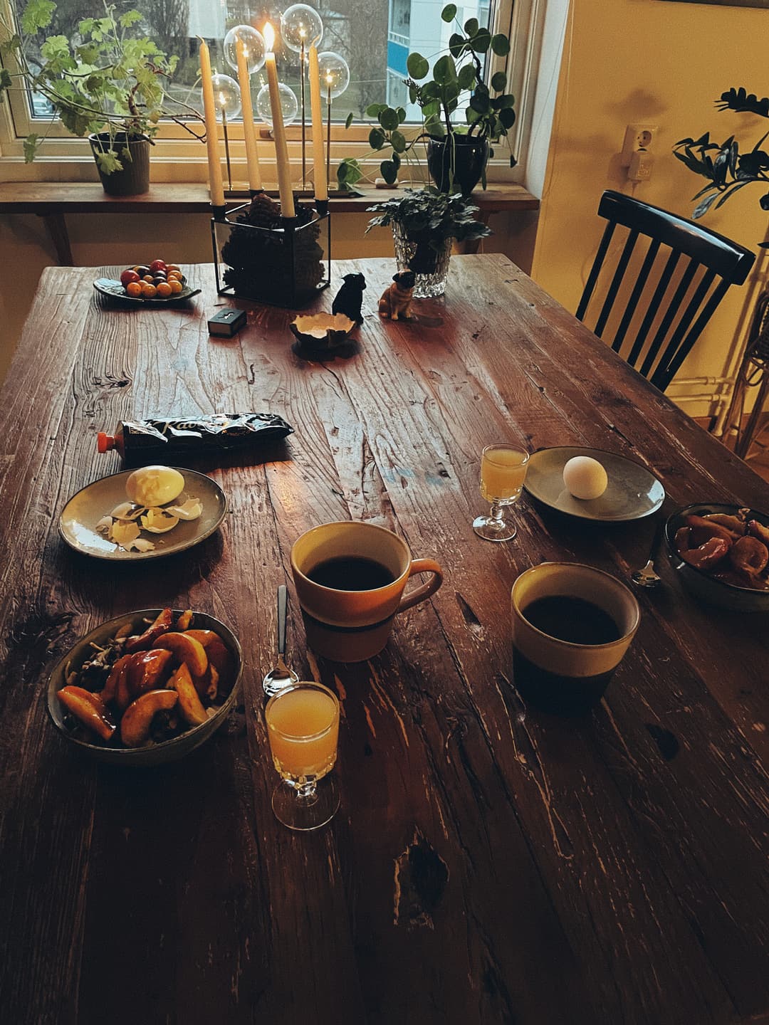 A rustic kitchen table. A lit candle makes for a cozy atmosphere. Coffee, eggs, energizing juice shot, and granola are served.