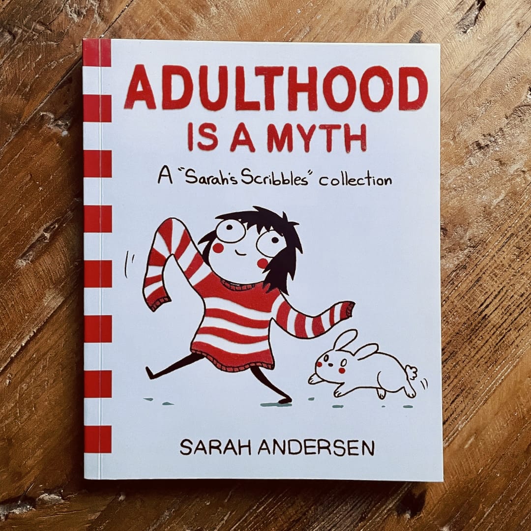 The cover of a book named Adulthood is a Myth by Sarah Andersen. A girl with messy hair wears an oversized, white sweater with red stripes. Or is it a red sweater with white stripes? The girl is followed by a white bunny.