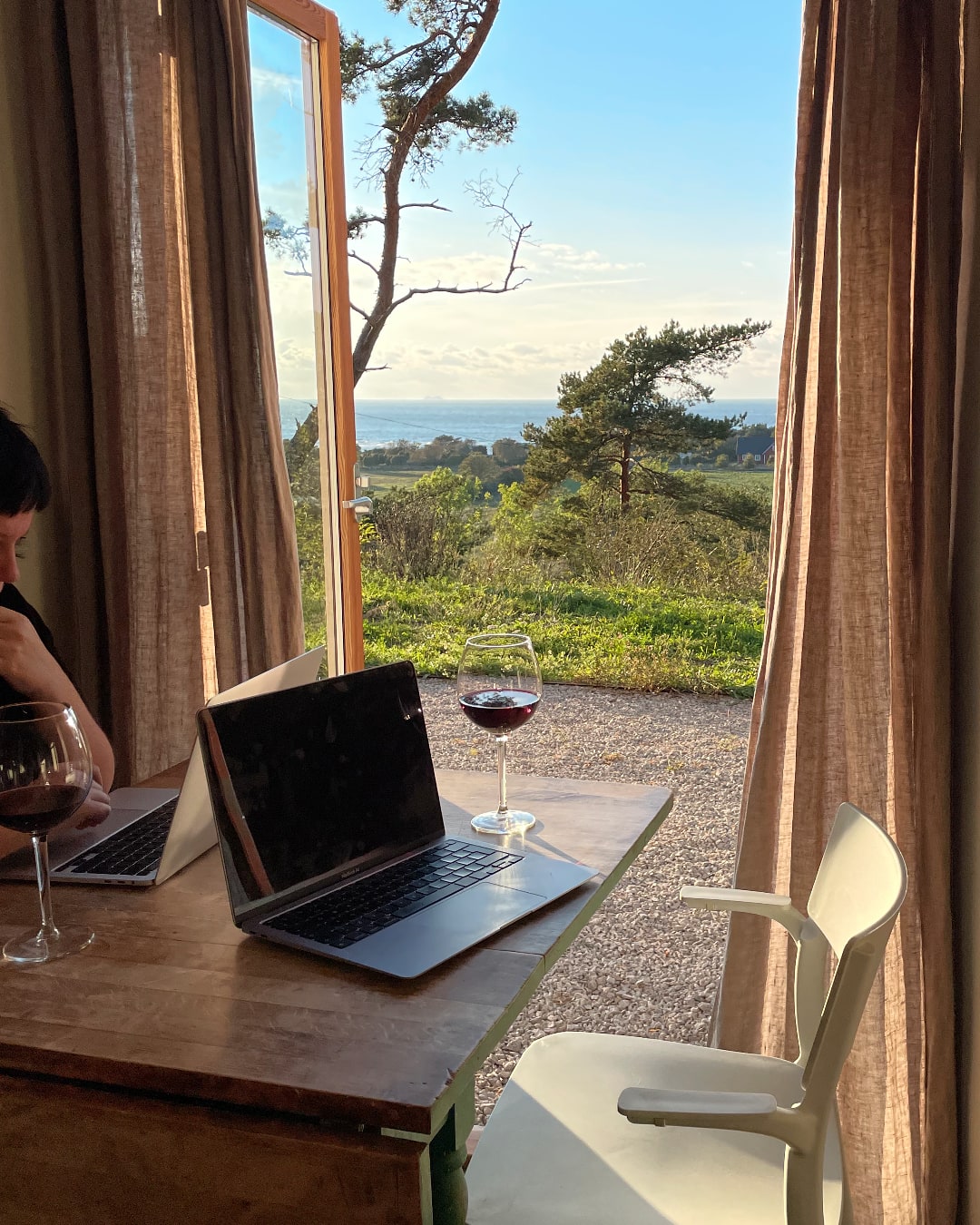 A spectacular view from an open door. A blue sky with clouds and the sea at a distance. Inside, a woman sits at a table working on a laptop accompanied by a glass of fine. Opposite her, there's another laptop, a glass of wine, and an empty chair. Maybe the photographer's seat?