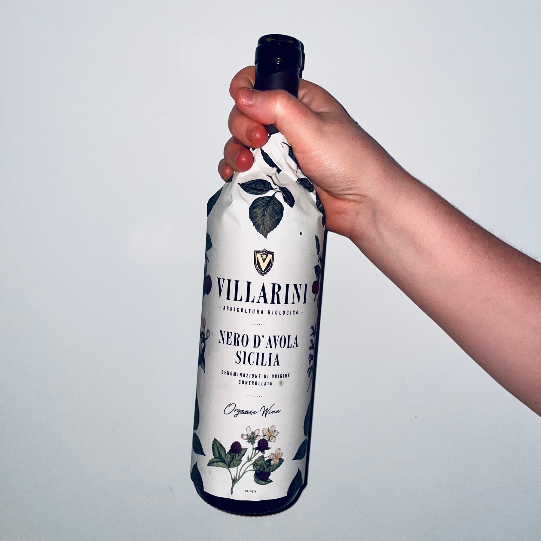 A hand holding an opened wine bottle. It's totally wrapped in paper, lovingly illustrated with leaves, that also act as the bottle's label. The label reads Villarini Nero d’Avola Sicilia, organic wine.