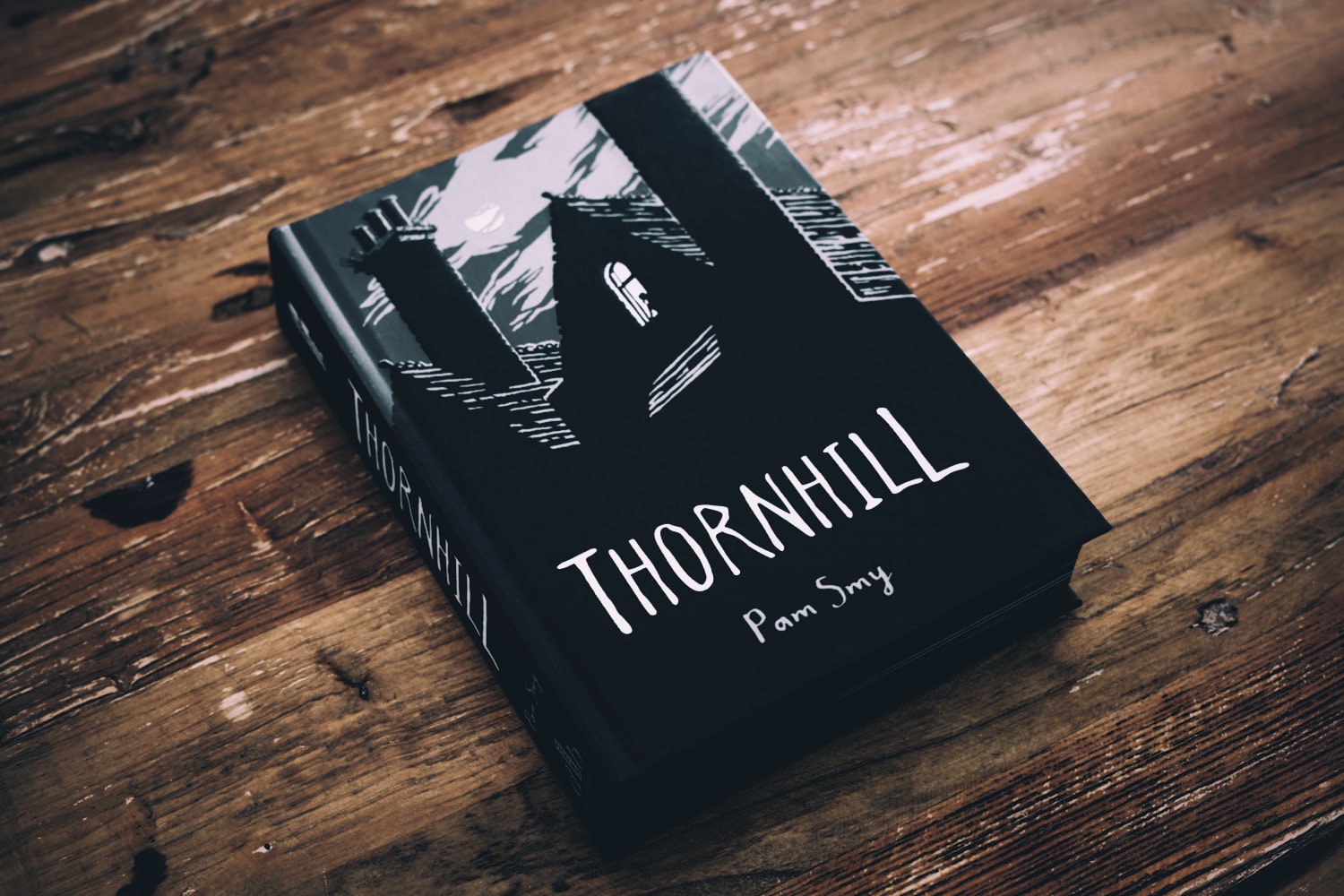 A pretty thick book, Thornhill by Pam Smy, lays on a wooden table. The cover is illustrated in black and white. The silhouette of a house with a prominent chimney on a moonlit night. A person, maybe a woman, looks out from a lit window. The title is handwritten in all caps.
