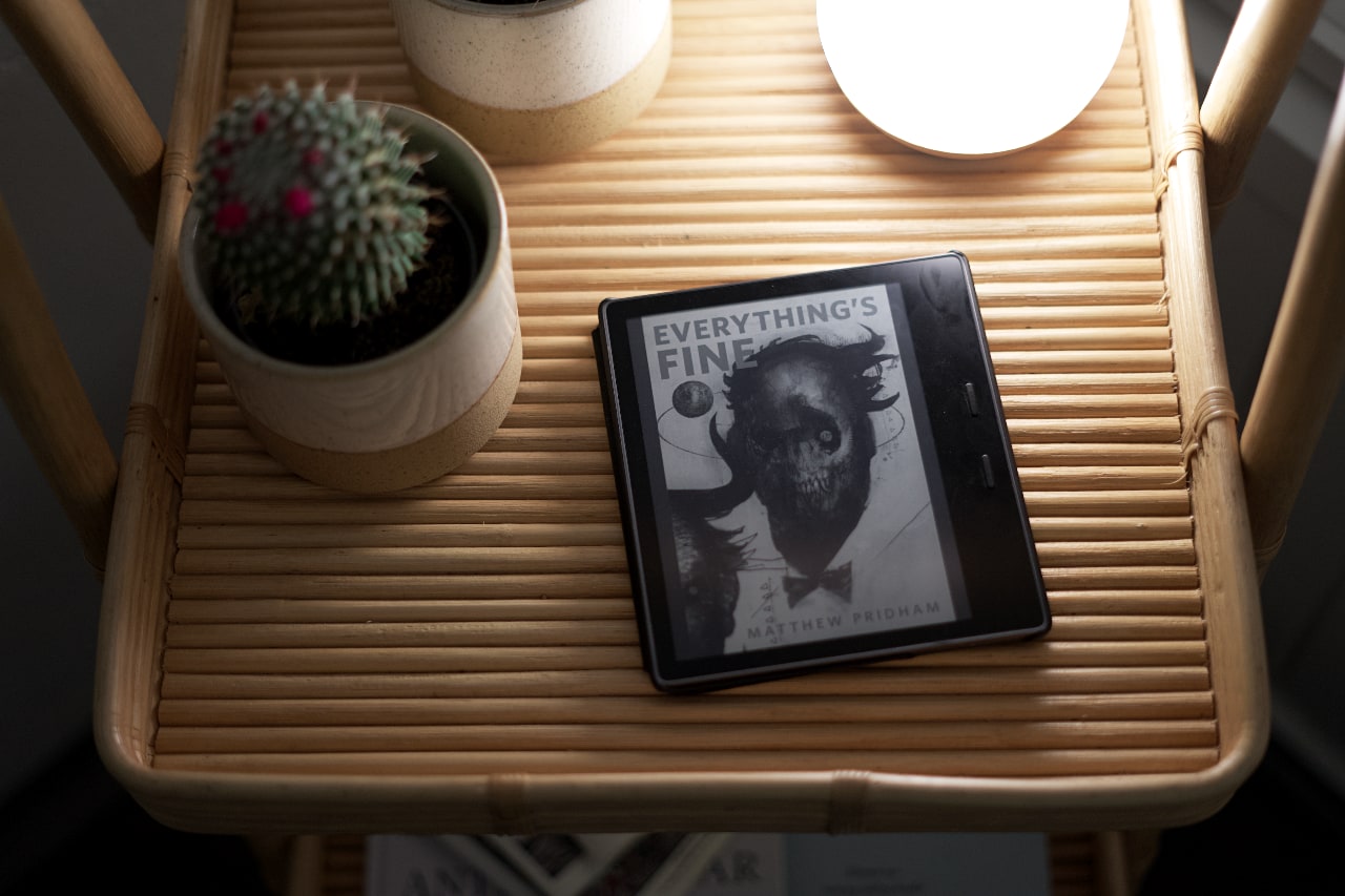 An e-reader on a tray, showing the cover of a short story, named Everything's Fine. It's illustrated with a skull wearing a tie. A tiny planet, maybe Earth, orbits around the head. The author's name at the bottom reads Matthew Pridham.