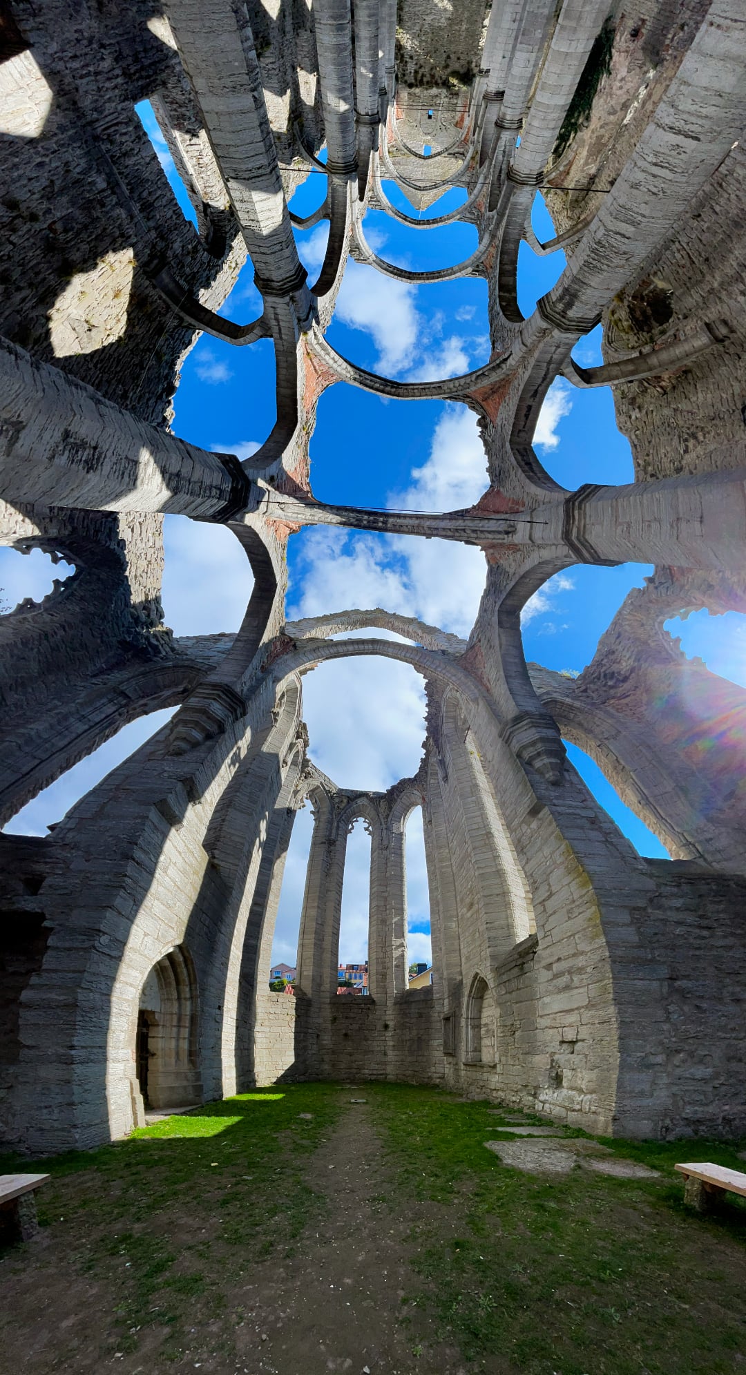 Interior of a church ruin. There's a blue sky and cumulus clouds. The photo is shot in panoramic mode with a visible warping defect due to the photographer lacking talent.