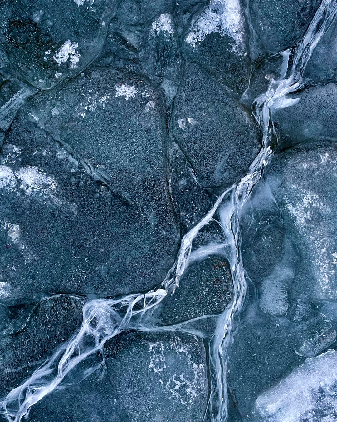 A top-down closeup of a frozen lake. There are cracks in the frosty ice, and the photo has a delicious blue tint over it.