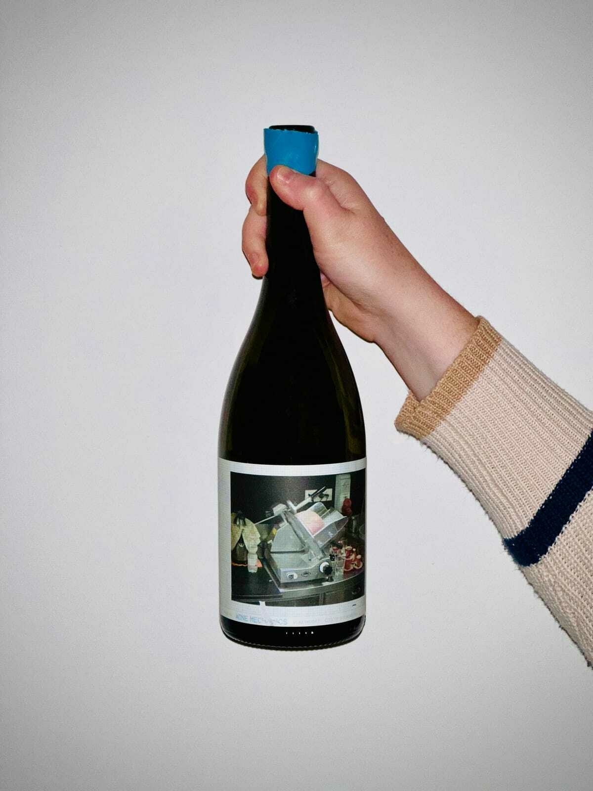 A wine bottle held up against a white wall. The label shows an illustration of a meat slicer. Wine Mechanics is barely readable.
