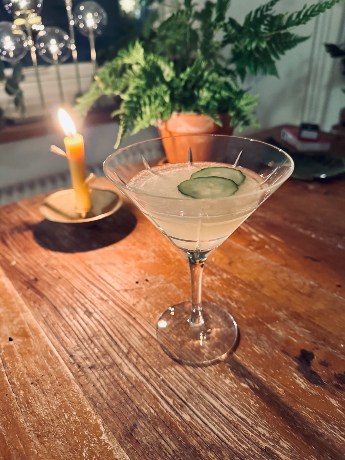 A cocktail with a hint of light green, adorned with a crisp cucumber slice, rests on a rustic wooden table. A softly glowing candle and a lively potted plant provide a cozy backdrop.