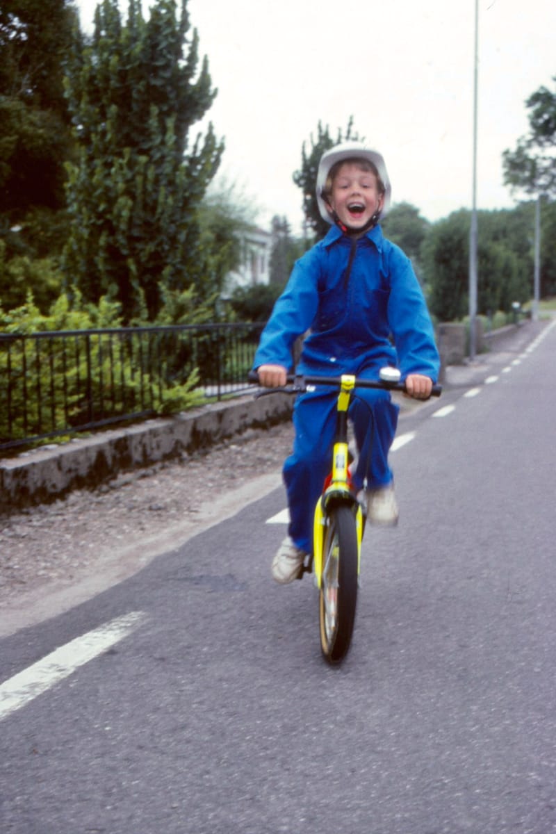 A kid in blue overalls, rocking a helmet, and flashing a big smile. Cruising straight towards the camera.