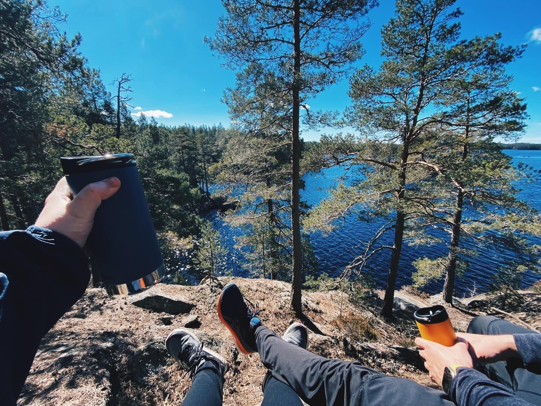 A couple sitting outdoors, sipping coffee from thermos mugs. Only their arms and legs are visible in the frame. The view over a lake is magnificent.