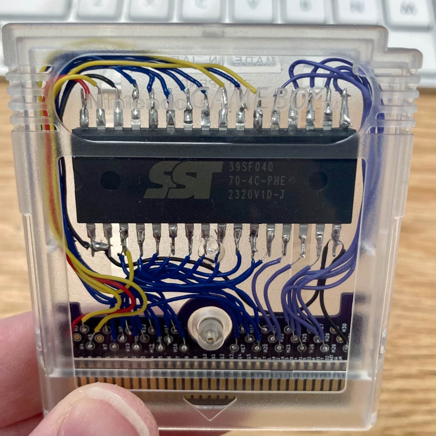 A translucent Game Boy cartridge, revealing the electronics inside. Numerous wires are soldered directly to the edge connector and flash chip—dead bug style. Messy but/and beautiful.