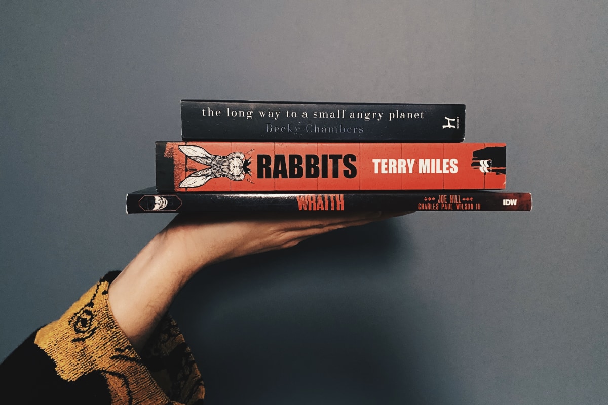A hand holding a pile of books. The long way to a small angry planet, by Becky Chambers, Rabbits, by Terry Miles, and Wraith, by Joe Hill.