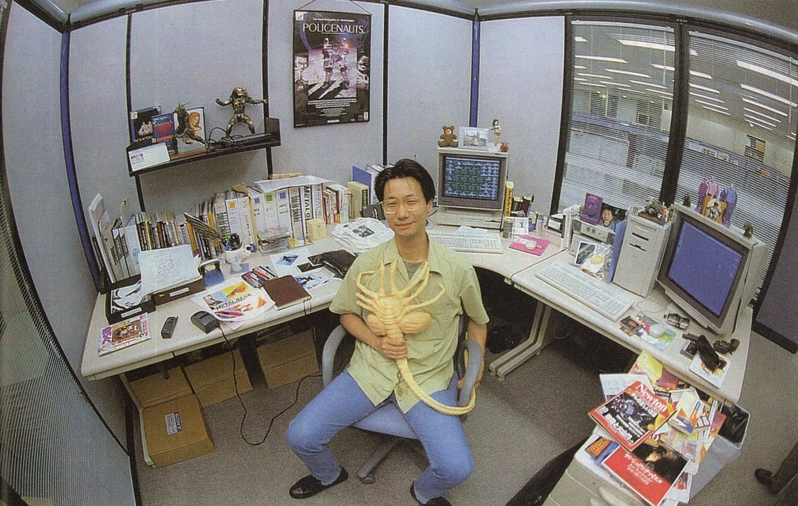 Hideo Kojima sitting turned away from his U-shaped desk in the late 90s. The desk has two CRT monitors and computers, and the rest of it is filled with stuff. Books, papers, magazines, CDs, photos, and toys. Kojima-san looks at the camera with a smile, holding a facehugger replica from the movie Alien.