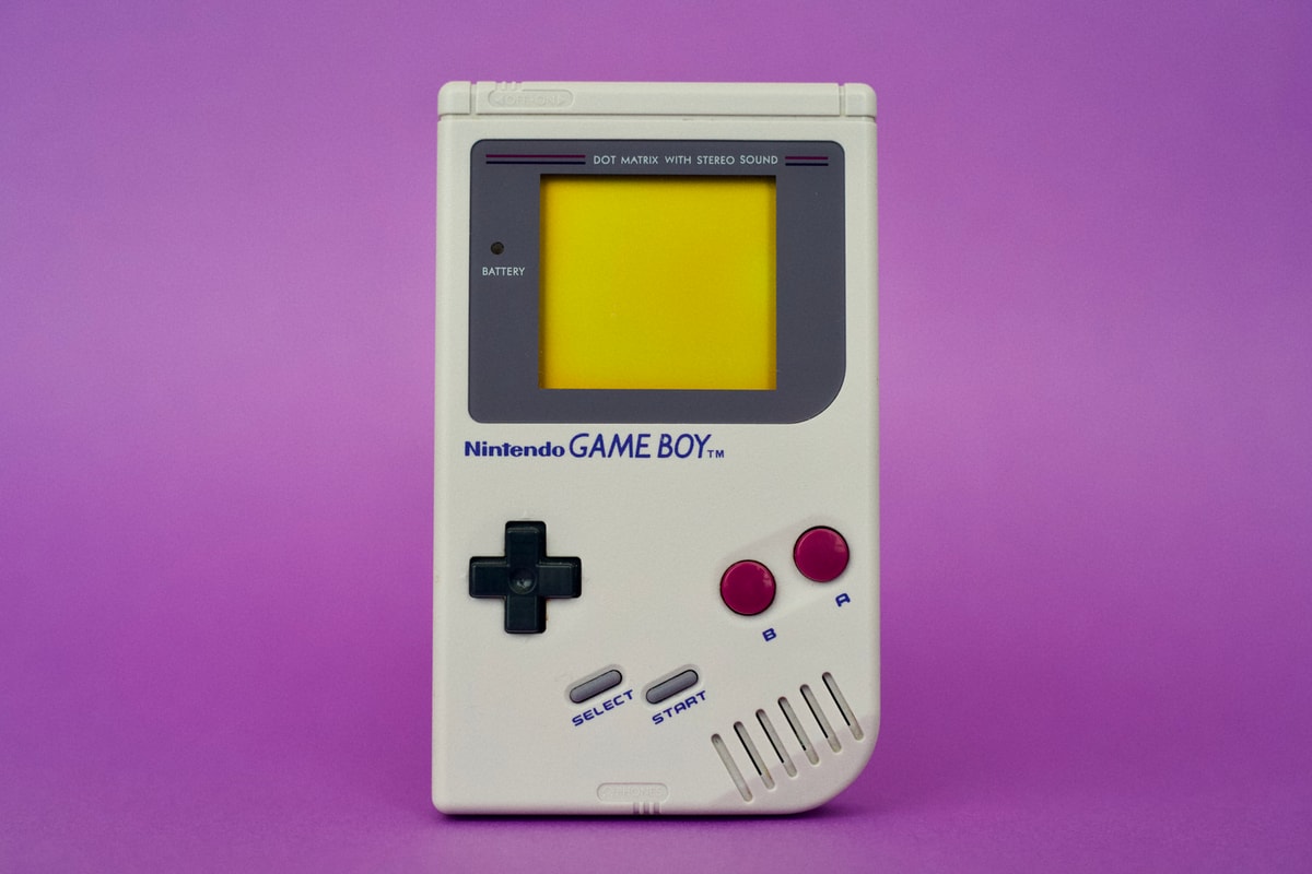 A Game Boy against a purpureus background. It looks like it was shot in a studio. The handheld is turned off.