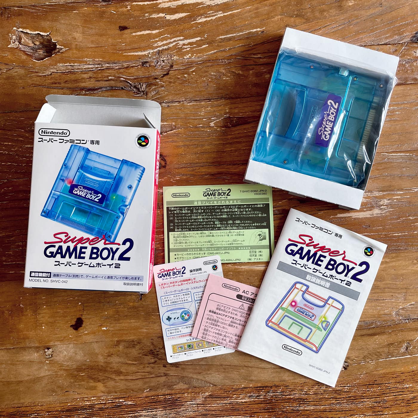 A Super Game Boy 2 accessory, original box, and instruction booklet lay on a wooden table. The cartridge is transparent blue, exposing the electronics inside.