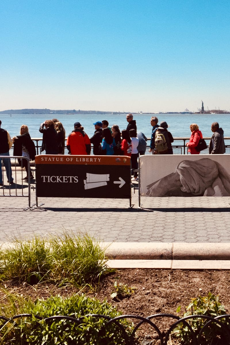 In the foreground, tourists cluster around a tickets sign. The Statue of Liberty, although distant, maintains a quiet but undeniable presence on the horizon.