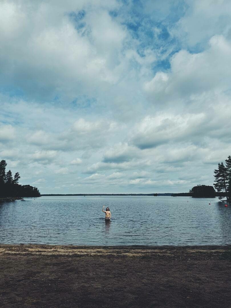 Me standing in the water, waving at the photographer, sandy shore in the foreground, surrounded by trees under a cloudy sky.