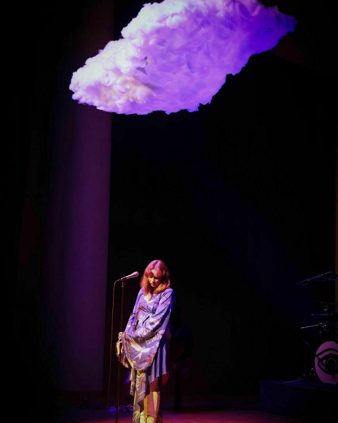 A woman (GERD) on stage with a microphone, looking down thoughtfully, with a large, illuminated cloud-like art installation floating above.