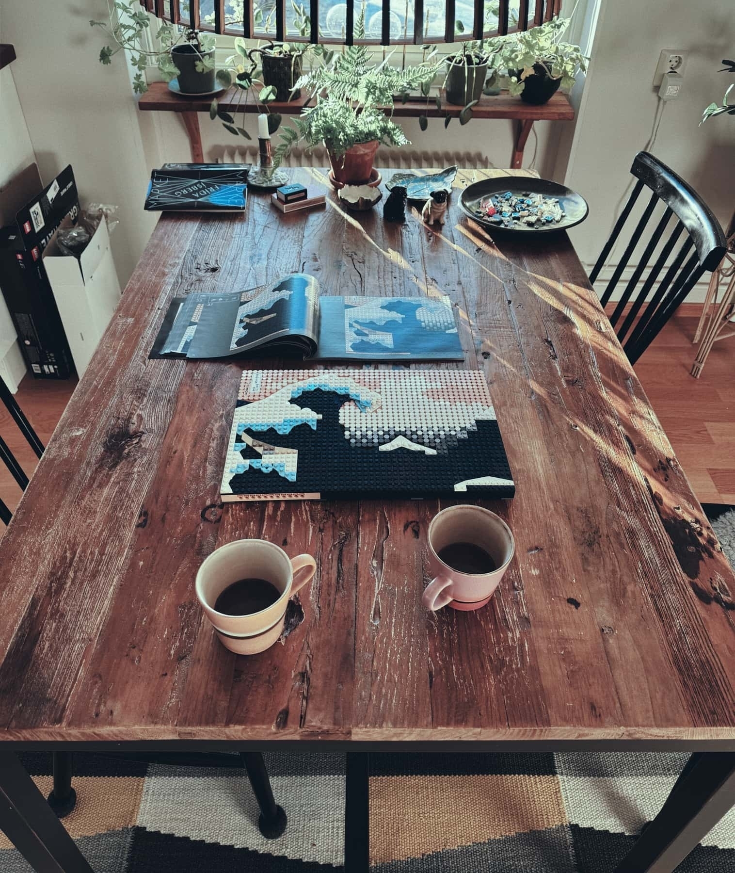 A wooden dining table with two cups of coffee and a partially completed Lego build of Hokusai’s The Great Wave.