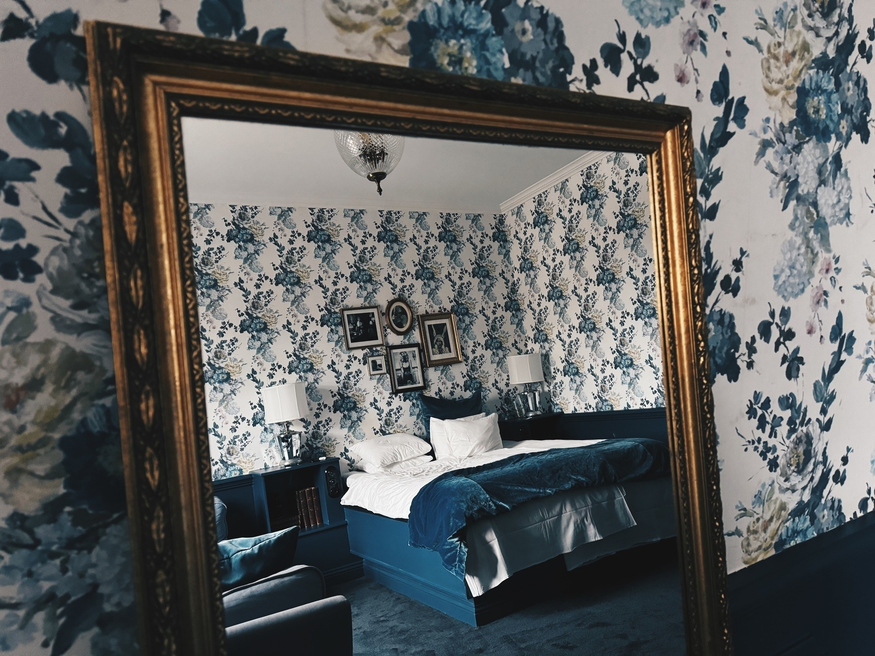 A golden mirror against a wall with a flower pattern. The mirror shows a cozy hotel bed.