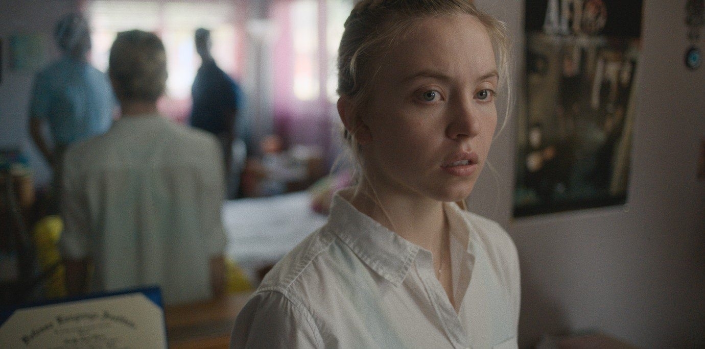 Still from the film, a close-up of a young woman, Sydney Sweeney, with a concerned expression looking off camera, with blurred figures in the background of a dimly lit room.
