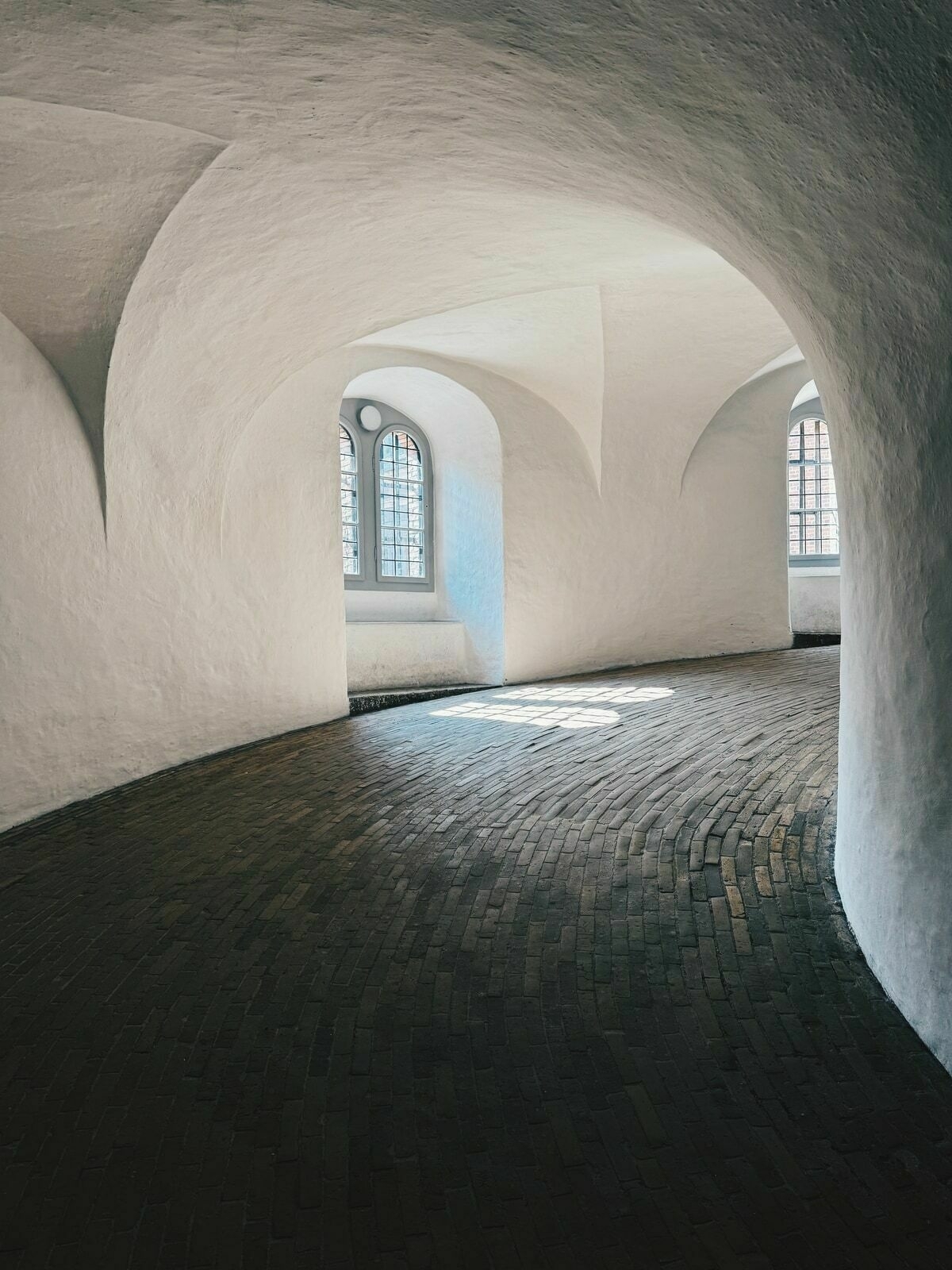 The tower’s vaulted interior with arched windows and cobblestone flooring. Sunlight filters through the windows, casting light patterns on the walls and floor. 