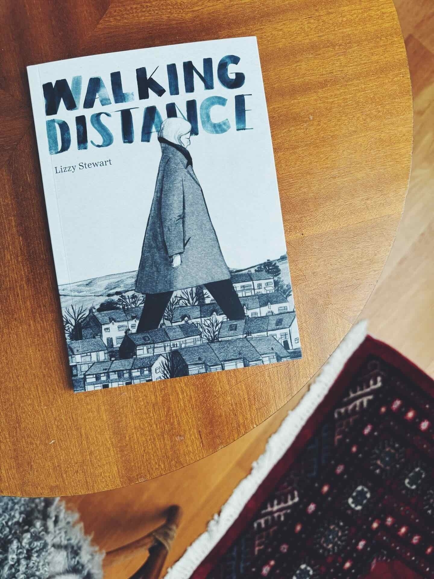 The book Walking Distance by Lizzy Stewart on a wooden table. The cover illustration depicts a large woman in a coat walking over a small town.